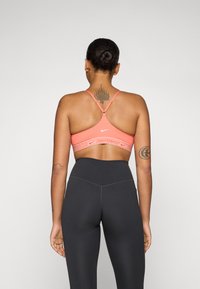 Sport bra u koralnoj boji s križnim naramenicama i širokim pojasom s "Nike Sportswear" logotipom. Nosimo ga s crnim tajicama visokog struka.