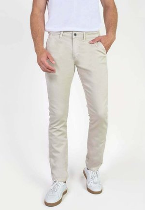 Homme portant un pantalon beige slim, des baskets blanches et une chemise blanche, une main dans la poche sur un fond uni.