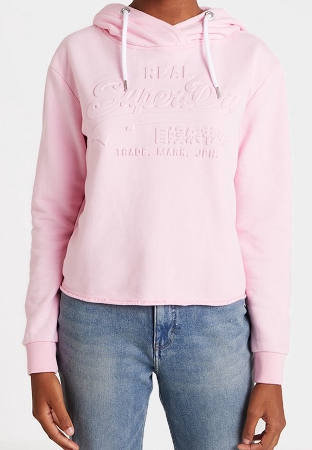 Personne portant un sweat à capuche rose clair avec du texte en relief et un jean bleu, faisant face à l'avant avec les mains détendues sur les côtés.