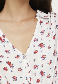 Blusa blanca con escote en V, con un pequeño cierre de botón y un estampado floral en rojo y azul. Tela ligera con una textura suave.