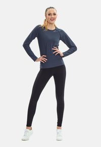 Long-sleeve navy athletic top with a round neckline, paired with black leggings and white sneakers. The fabric appears smooth and flexible.