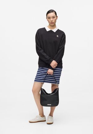 Vrouw in een zwart shirt met lange mouwen en witte kraag, blauw-wit gestreepte rok, zwarte handtas vasthoudend, witte sneakers dragend, staand op een witte achtergrond.