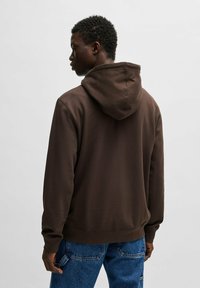 Felpa a capuccio marrone con una texture morbida e polsini a costine, indossata con jeans in denim blu, che mostra un design casual dalla schiena.
