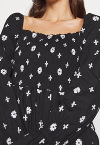 Femme portant une robe noire avec des motifs de croix blanche et de fleurs, avec un décolleté carré et des manches longues bouffantes.