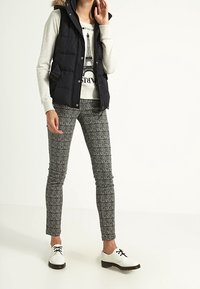 Kobieta ubrana w czarną kamizelkę puchową, jasny sweter z nadrukiem Wieży Eiffla, teksturowane szare spodnie i białe buty na sznurówki stoi na białej podłodze.