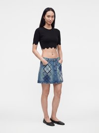 Νεαρή γυναίκα φορούσε μαύρο crop top με κυματιστή άκρη, μπλε τζιν φούστα με σχέδια argyle και μαύρα φλατ παπούτσια, στέκεται με τα χέρια στις τσέπες.