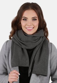 Grau gestrickter Schal aus einem weichen, strukturierten Stoff, um den Hals drapiert mit nahtlosem Design und dezentem Branding-Detail am Ende.
