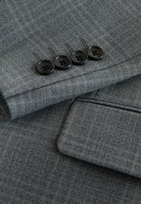 Nahaufnahme eines grauen Stoff-Sakko mit glatter Textur, vier schwarzen Knöpfen am Ärmel und einem sauber vernähten Taschendetail.