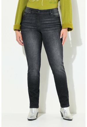 Frau trägt schwarze Skinny-Jeans mit hoher Taille, hellgrünes langärmliges Oberteil mit hochgekrempelten Ärmeln und silberne Stiefeletten, steht vor weißer Wand.