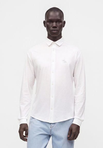 Modèle masculin portant une chemise blanche unie à boutons avec col et un jean bleu clair, debout les bras détendus devant un fond blanc.