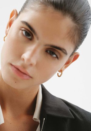 Pendiente de aro dorado retorcido exhibido en un modelo. Primer plano que muestra la superficie texturizada y la piel suave contra un fondo neutral.