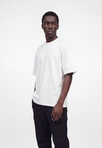 Giovane uomo dalla pelle scura che indossa una semplice t-shirt bianca oversize e pantaloni cargo neri, in piedi davanti a uno sfondo bianco.