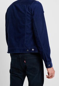 Marinblå manchesterjacka med rak passform, tryckknappar och texturerat tyg, kombinerad med mörka jeans i denim med synlig märkesetikett.