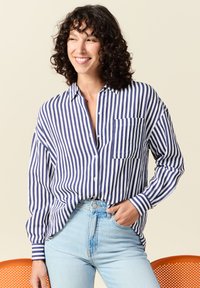 Camicia a righe blu e bianche con bottoni, maniche lunghe, taschino sul petto e vestibilità rilassata, abbinata a jeans chiari a vita alta.