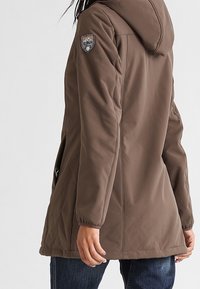 Veste marron à capuche avec fermeture zippée, ornée d'un patch sur l'épaule gauche, et de longueur jusqu'à la taille avec deux poches latérales.