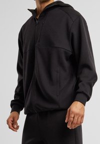 Sudadera con cremallera negra, fabricada en un tejido suave. Presenta una capucha, dos bolsillos laterales y puños acanalados. Textura lisa con detalles de costura mínimos.