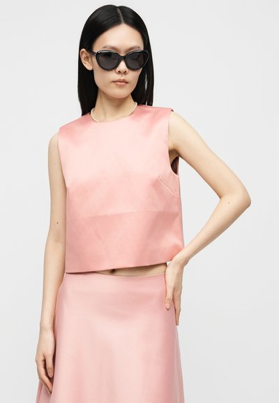 Γυναίκα που φοράει μαύρα γυαλιά ηλίου cat-eye, ρόζ σατέν τοπ χωρίς μανίκια και ταιριαστή ρόζ φούστα, ποζάροντας με το ένα χέρι στη μέση.