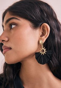 Gold sun-shaped earrings with a black fringe design, featuring a circular cutout. Earrings hang from a stud and have a textured finish.