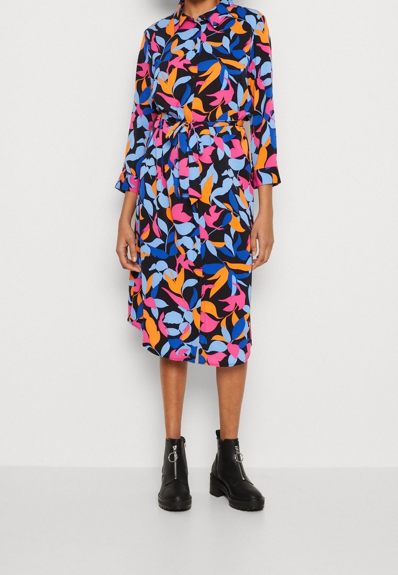 Personne portant une robe mi-longue ceinturée avec un motif abstrait de feuilles colorées en bleu, rose, orange et noir, et des bottines noires.