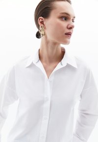 Witte button-up shirt met een kraag en een gestreept binnendetail. Model draagt gedurfde gouden oorbellen in de vorm van bladeren. Glad materiaal, ontspannen pasvorm.