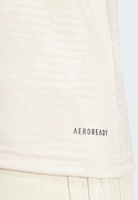 T-shirt beige clair avec un motif texturé de formes géométriques blanches. Présente le logo "AEROREADY" en noir près de l'ourlet. Tissu doux.