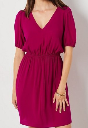 Femme portant une robe magenta mi-longue avec un col en V, des manches bouffantes, une taille élastique et des bracelets dorés au poignet droit.