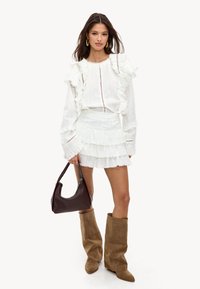 Witte getextureerde top met ruffles, witte gelaagde rok, bruine suède knielange laarzen en een donkerbruine schoudertas met een kralenhandvat.