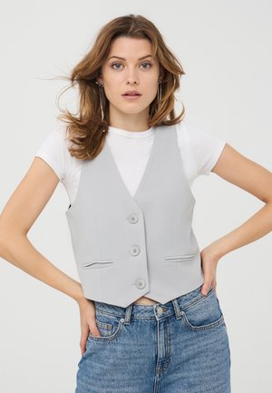 Gilet grigio chiaro con un design aderente, caratterizzato da un collo a V, cinque bottoni e due tasche frontali. Indossato sopra una maglietta bianca e jeans in denim.