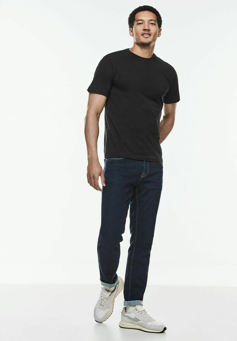 Zwarte katoen T-shirt met een ronde hals, gecombineerd met donkerblauwe jeans en lichtgrijze sneakers. Model staat in een ontspannen houding.