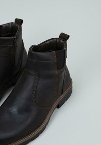 Un par de botas de cuero marrón oscuro hasta el tobillo con paneles elásticos laterales, tiradores y tacones bajos sobre un fondo gris claro.