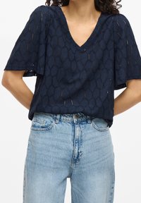 Woman wearing a navy blue textured V-neck short sleeve blouse tucked into light blue high-waisted jeans with hands behind her back.