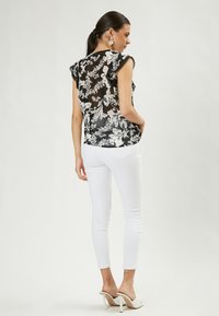 Schwarze durchsichtige Bluse mit weißem Blumenmuster, Rüscheärmeln und lockerer Passform, kombiniert mit weißen Skinny-Jeans und offenen Pumps.