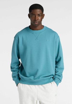 Young man wearing a teal sweatshirt and light gray sweatpants, standing with hands in pockets against a plain light background.