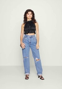 Jovem mulher com cabelo cacheado, vestindo um top cropped preto sem mangas, jeans azuis rasgados, sapatos de salto pretos e uma pequena mala de ombro.