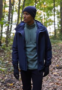 Veste imperméable navy avec une capuche, superposée sur une chemise en polaire verte. Gants noirs, debout au milieu du feuillage d'automne dans une forêt.