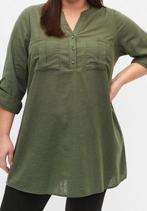 Femme portant une tunique verte olive à manches longues avec revers retroussés, col en V boutonné, et deux poches poitrine, associée à un pantalon noir.