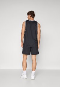 Top sin mangas atlético negro con ribetes a rayas blancas, combinado con pantalones cortos de malla negros. Lleva zapatillas blancas y calcetines blancos visibles.