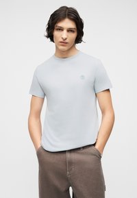 Νεαρό άτομο με μαλλιά μεσαίου μήκους σκουρόχρωμα, που φοράει ανοιχτό μπλε κοντομάνικο πουκάμισο και καφέ παντελόνι, με τα χέρια στις τσέπες, ουδέτερο φόντο.