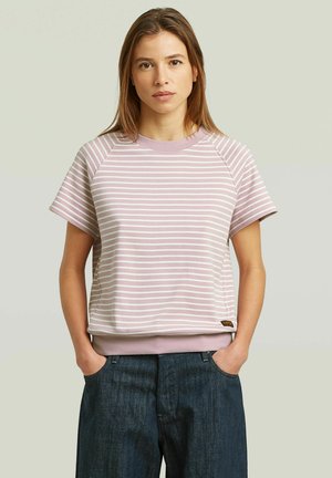 Vrouw met recht bruin haar, gekleed in een lichtpaars met witte gestreepte top met korte mouwen en donkerblauwe jeans, staand met handen in de zakken.