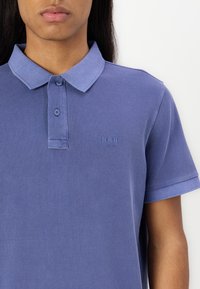 Short-sleeve, collared polo shirt in light blue cotton. Features two-button placket and tonal embroidered logo on the left chest.