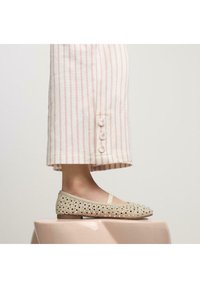 Ballet flat beige con design perforato e dettagli in strass. Punta arrotondata e tacco basso, abbinati a pantaloni a righe con dettagli a bottoni.