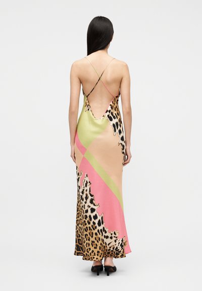Vrouw draagt een lange, rugloze slipjurk met dunne gekruiste bandjes, met een luipaardprint en roze en groene diagonale kleurvlakken.