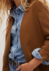 Blazer marron superposé sur une chemise en denim bleu clair, avec un fermeture à boutons et des manches retroussées, accompagné d'une ceinture noire et d'une texture en denim.