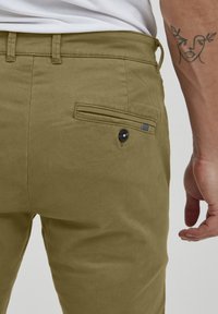 Chinos vert olive avec une texture lisse, comportant une poche arrière avec un bouton noir et un petit accent rayé.