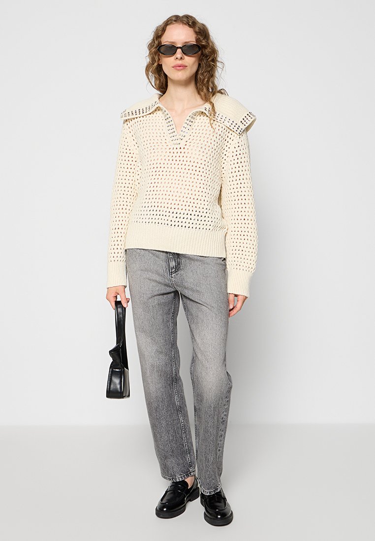 Strickpullover in cremefarben mit Lochmuster und oversized Kragen. Kombiniert mit grauen Geradepassform-Jeans, schwarzen Schuhen und einer kleinen Tasche.