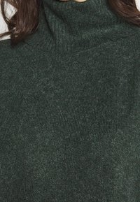 Ciemnozielony sweter z golfem wykonany z miękkiej, teksturowanej dzianiny z prążkowanym dekoltem. Powierzchnia wydaje się puszysta z drobnymi włóknami.