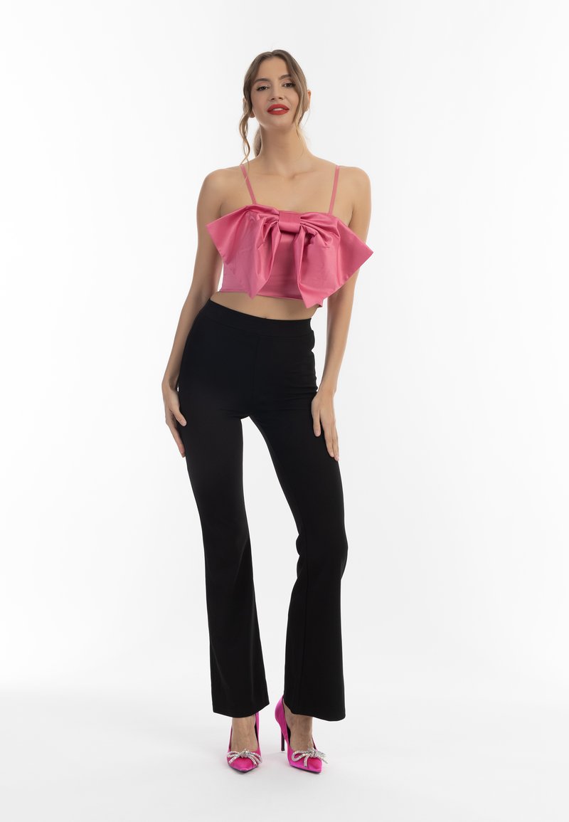 Roze cropped top met oversized strikdetail, gecombineerd met zwarte flared broeken. Model draagt roze hoge hakken met decoratieve accenten.