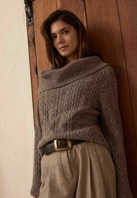 Femme aux cheveux bruns portant un pull en maille torsadée marron avec un grand col et un pantalon beige taille haute avec une ceinture noire, debout contre une porte en bois.