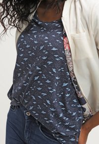 Mörkblå kortärmad skjorta med ljusblå fågelmönster, kombinerad med en krämfärgad jacka och mörka jeans. Mjuk, avslappnad passform.