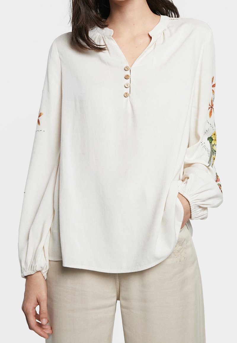 Kvinna klädd i en löst sittande krämfärgad blus med träknappar och blommönster på ärmarna, matchat med beige byxor, med handen i fickan.